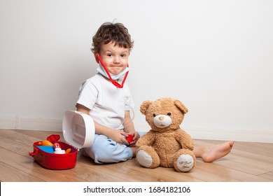 A Small Child, A Boy In A Medical Mask With A Stethoscope On His Neck Plays A Doctor With A Children's Medicine Cabinet, Gives A Teddy Bear An Injection And Measures The Temperature.