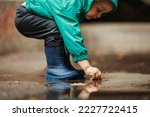 A small child boy wearing blue rubber boots and a jacket jumping through puddles and playing outside on a rainy day. Having fun outdoors during a bad weather. Hands in a water. Copy space.