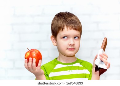 Small Child Chooses Chocolate Or Apple On A Light Background In The Studio.