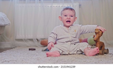 A Small Child Is Crying Sitting On The Carpet In The Nursery. Toddler Baby Boy Is Naughty On The Floor With Toys, Age Eight Months