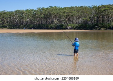 A small child fishing with a fishing rod, standing in still, reflective water with golden sand and trees in the background. - Shutterstock ID 2254888189