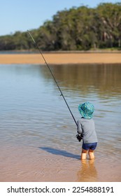 A small child fishing with a fishing rod, standing in still, reflective water with golden sand and trees in the background. - Shutterstock ID 2254888191