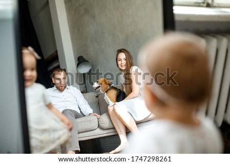A small child girl stands near a large mirror and looks at her parents and a dog. In reflection parents. Mom, dad and beagle dog. A happy family. Parents are in focus