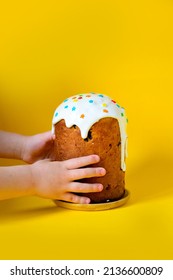 A small child is holding an Easter cake with white meringue and multicolored decor on a yellow background. The concept of the traditional Easter holiday