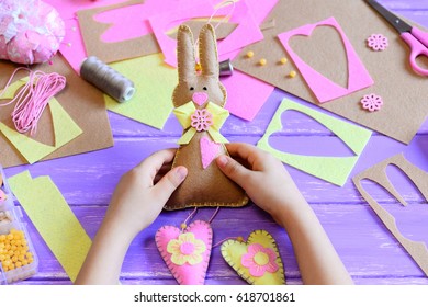 Small Child Holds A Felt Easter Bunny In His Hands. Child Made A Felt Cute Bunny With Hearts For Easter. Tools And Materials For Art Creativity On A Wooden Table. Simple Spring Crafts Project For Kids