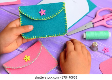 Small Child Holds A Felt Purse In His Hand. Child Shows A Felt Purse. Handcraft Supplies On A Wooden Table. Funny And Easy Hand Sewing Project For Kids 