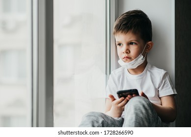 A Small Child In A Medical Mask Sits Quarantined At Home On A Window With A Phone In His Hands.Prevention Of Coronavirus And Covid - 19