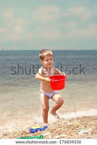 A small child on the beach.Boy carrying water from the sea in a red bucket. In all growth. Vertical photos.
