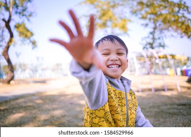 Small Child Playing In A Park Spreading Out His Hand Forward Reaching To Camera, Little Kid Smiling With White Teeth And Raising His Arm Toward Camera With Happy Face During Beautiful Sunny Day