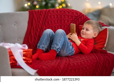 Small Child Plays With Technical Device Alone. Boy Looks At Smartphone Screen While Lying On Couch At Christmas. Kids Addicted To Smartphone Or Game Concept. Playing With Smartphone Addiction.