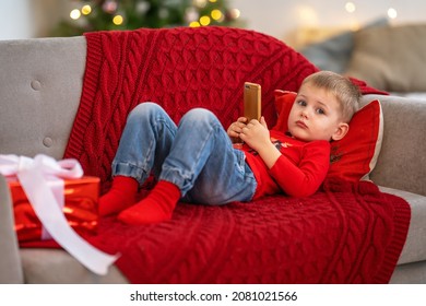 Small Child Plays With Technical Device Alone. Boy Looks At Smartphone Screen While Lying On Couch At Christmas. Kids Addicted To Smartphone Or Game Concept. Playing With Smartphone Addiction.