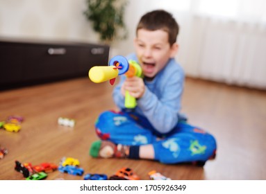 A Small Child A Preschool Boy Is Playing A Shooting Game With A Toy Gun On The Background Of A Bright Game Room With Toys. A Foam Bullet Or Rocket Flies Out Of A Toy Machine Gun.