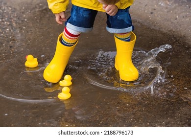 A small child in rainbow socks, yellow rubber boots and a jacket jumps through puddles and plays with yellow rubber ducks. A picture of summer and autumn holidays. A child in the rain. - Shutterstock ID 2188796173