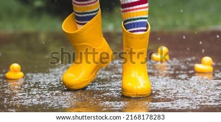 A small child in rainbow-colored socks and yellow rubber boots jumps through puddles and plays with yellow rubber ducks. Legs close-up. A picture of summer and autumn holidays. A child in the rain.