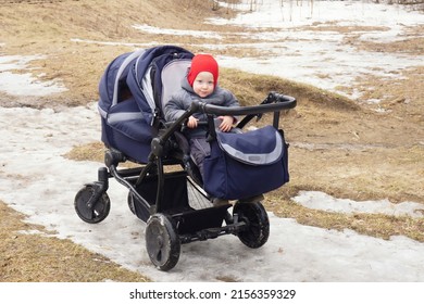 Small Child In Red Cap In Double Blue Pram Looks At The Camera And Smiles. Cloudy, Early Spring, Melting Snow, Last Year's Grass