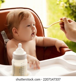 Small Child Sits On A Chair And Eating With Spoon. Little Smiling Girl Sits In Baby-chair And Have A Breakfast. Baby With Big Eyes And Red Curly Hair Eats At A Table. Summer Vacation Concept.