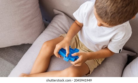 A Small Child Is Sitting On The Sofa And Holding A Pop It Toy. Portrait Of Concentrated Cute Little Boy Playing Popit Toy. Antistress Toy For Children. Leisure For Kids. Concept Of Mental Health, Calm