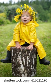 A small child in a yellow raincoat and a buttercup wreath sits on a stump.