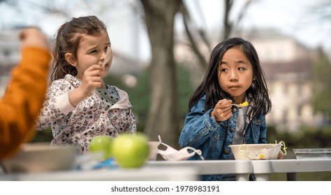 Small Children Eating Lunch Outdoors In City Park, Learning Group Education And Coronavirus Concept.