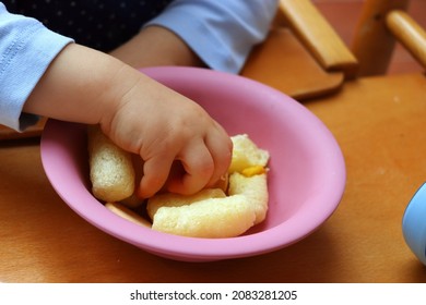 A small child's hand takes a snack from a pink bowl placed on a wooden children's table. Extruded baby cornflakes.