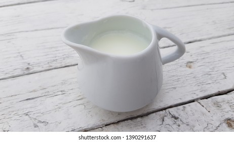 Small China Milk Jug Full With Milk On White Distressed Wooden Table. White Crockery Closeup On Shabby Wood. Kitchen Faience Utensil. All White Minimal Still Life. Milk For Morning Cup Of Coffee.