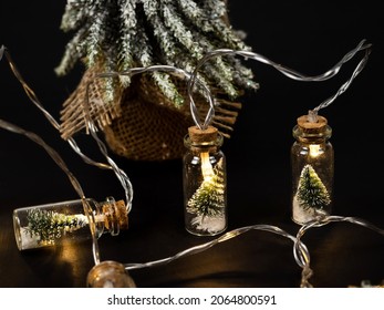 Small Christmas tree on a black background. Glowing lights decorating glass jars with corks, inside are small green Christmas trees on artificial snow. Christmas composition. Christmas, New Year conce - Powered by Shutterstock