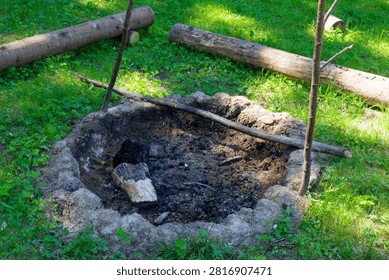 Small circular fire pit dug in the ground with charred wood and embers surrounded by grassy clearing, rustic campsite equipment prepared for an outdoor adventure in the green woods