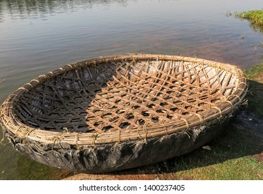 A Small Circular Lightweight Boat Or Coracle Constructed Of Natural Materials Woven Together In An Intricate Pattern To Give Strength And Buoyancy. The Craft Is Resting On A Grass Bank Beside A River.