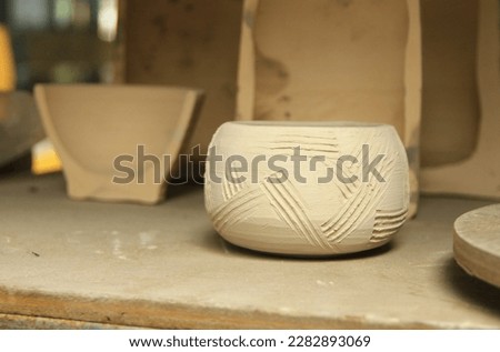 A small clay bowl made by ceramic throwing machine. Decorated with straight lines from incising technique and has a rough texture. Air-dried and unburnt. Displayed on a wood shelf in ceramic workshop.