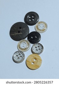 A small collection of assorted sewing buttons in different sizes, colors, and designs on a white surface. The set includes black, dark brown, cream, and translucent white buttons with two hole and four hole styles. Some have gold glitter trim, wood grain texture, or a silver rim with cutout patterns for a mixed craft and tailoring feel.