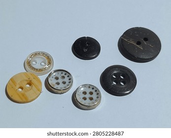 A small collection of assorted sewing buttons in different sizes, colors, and designs on a white surface. The set includes black, dark brown, cream, and translucent white buttons with two hole and four hole styles. Some have gold glitter trim, wood grain texture, or a silver rim with cutout patterns for a mixed craft and tailoring feel.