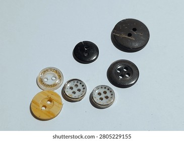 A small collection of assorted sewing buttons in different sizes, colors, and designs on a white surface. The set includes black, dark brown, cream, and translucent white buttons with two hole and four hole styles. Some have gold glitter trim, wood grain texture, or a silver rim with cutout patterns for a mixed craft and tailoring feel.
