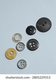 A small collection of assorted sewing buttons in different sizes, colors, and designs on a white surface. The set includes black, dark brown, cream, and translucent white buttons with two hole and four hole styles. Some have gold glitter trim, wood grain texture, or a silver rim with cutout patterns for a mixed craft and tailoring feel.
