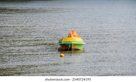 small, colorful boat floats on calm water. The boat is painted in bright shades of green, yellow, and orange. A yellow buoy is attached to the boat by a rope. - Powered by Shutterstock