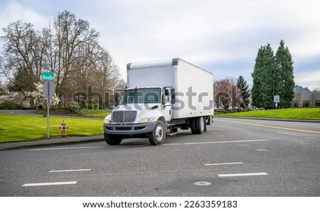 Small compact bonnet white semi truck rig with cube box trailer transporting commercial cargo driving on the street of urban city in spring time with blooming trees
