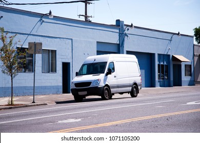 Small Compact White Minivan For Commercial Transportation And Small Business - Convenient And Practical Vehicle Standing On Urban City Street With Warehouse Buildings