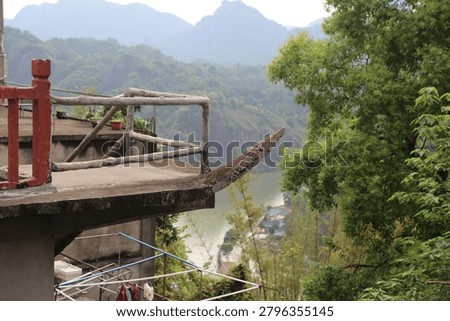 A small concrete balcony projects from a hillside building with rough branch-style railings and a curved slab edge. Green trees, a muted lake, distant mountains, and laundry lines below create a layered outdoor scene.