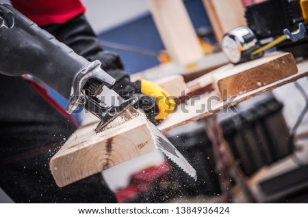 Small Construction Project. Men Cutting Piece of Wood Board Using Reciprocating Saw. Construction Site Power Equipment.