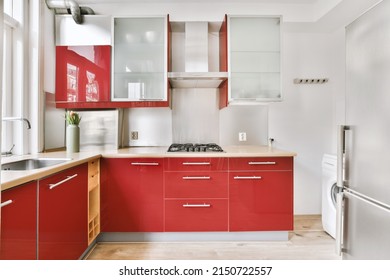 Small Corner Kitchen With Sink Under Window And Red Furniture With Transparent Cupboards With Stove, Fridge And Washing Machine In Urban Apartment