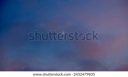 A small crescent moon against a blue sky. View of a crescent moon through clouds in a blue-violet sky during the day.                               