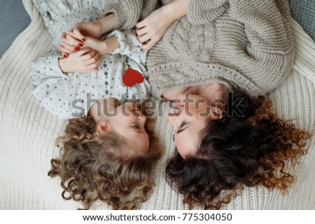 Small curly girl and her mother lie together in bed, looks at each other with great love, enjoy togetherness. View from above of affectionate mother and daughter. Children and parents concept