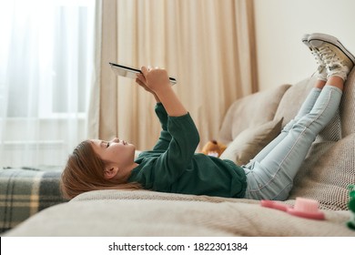 A Small Cute Girl Playing Games On A Tablet Lying On A Sofa With Her Feet Up And Gumshoes On In A Big Room Being Alone At Home. Children And Modern Technologies