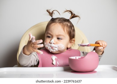 Small cute kid brunette girl with two tails tasting by herself with a spoon and dirty hand the greek yogurt, sitting in baby highchair with messy face and bib, self-feeding concept - Shutterstock ID 2239761637