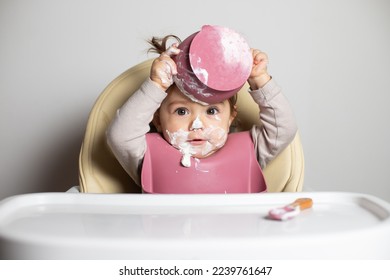 Small cute kid brunette girl with two tails eating by herself the greek yogurt and raising the dirty bowl up, sitting in baby highchair with messy face and bib, self-feeding concept - Shutterstock ID 2239761647