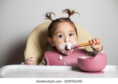 Small cute little kid brunette caucasian girl with two tails tasting and enjoying by herself with a spoon the greek yogurt sitting in baby chair with messy face; self-feeding concept - Shutterstock ID 2239761545