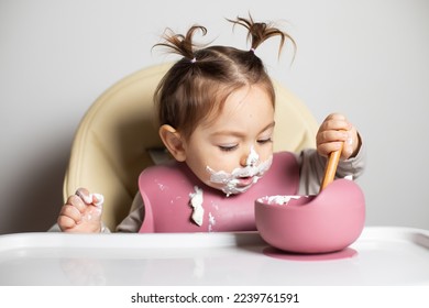 Small cute little kid brunette caucasian girl with two tails tasting by herself with a spoon the greek yogurt sitting in baby chair with messy face and bib; self-feeding concept - Shutterstock ID 2239761591