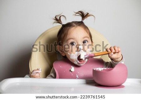 Small cute little kid brunette caucasian girl with two tails tasting and enjoying by herself with a spoon the greek yogurt sitting in baby chair with messy face; self-feeding concept