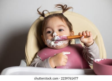 Small cute little toddler brunette caucasian girl with two tails eating by herself with a spoon the greek yogurt sitting in baby chair; self-feeding concept - Shutterstock ID 2239761477