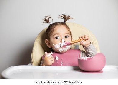 Small cute little toddler brunette caucasian girl with two tails tasting and enjoying by herself with a spoon the greek yogurt sitting in baby chair with messy face; self-feeding concept - Shutterstock ID 2239761537