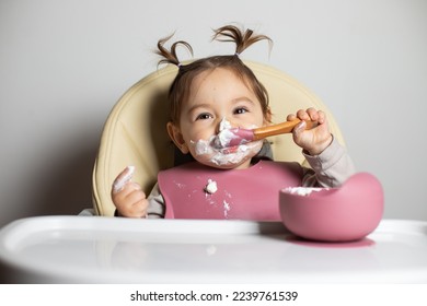 Small cute little toddler brunette caucasian girl with two tails tasting and enjoying by herself with a spoon the greek yogurt sitting in baby chair with messy face; self-feeding concept - Shutterstock ID 2239761539